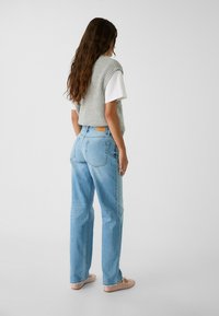 Vaqueros rectos de color azul claro con bolsillos traseros y un parche de cuero marrón, combinados con un chaleco de punto gris sobre una camiseta blanca.