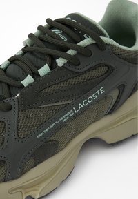 Lacoste sneaker in dark green mesh and suede, featuring a textured design, contrasting light green accents, and a beige rubber sole.
