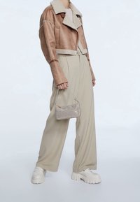 Afgeknipte bruine shearlingjas met een beige kraag, gecombineerd met lichtbeige wijde broek en witte platformsneakers, met een geweven mini tas in de hand.