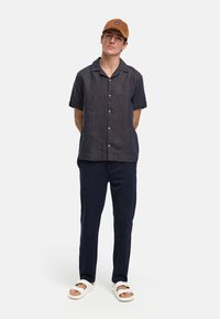 Camisa de lino de manga corta en azul marino con botones, combinada con pantalones oscuros y sandalias blancas. La gorra marrón presenta un logotipo. Diseño sencillo.