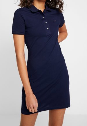 Granatowa sukienka polo wykonana z bawełny, z kołnierzykiem, listwą na guziki i dopasowanym krojem. Logo Lacoste w tym samym kolorze na wysokości klatki piersiowej.