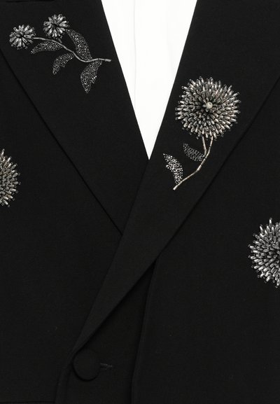 Schwarzer Blazer mit silbernen Perlenblüten und Blattan embellierungen an den Revers und vorderen Panele, getragen über einem weißen Kleidungsstück.