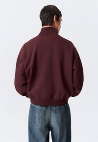 Sweatshirt grená com gola alta, punhos e barra canelados. Usada com jeans azuis com bolsos traseiros. Tecido macio, corte descontraído.