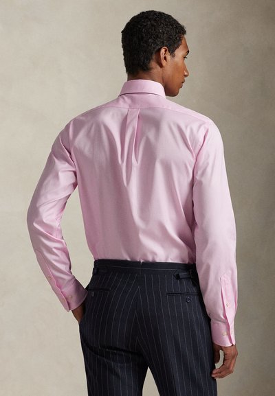 Férfi rózsaszín hosszú ujjú inget és sötétkék tűcsíkos nadrágot visel, háttal áll, jobb kezét a csípőjén nyugtatva.