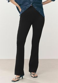 Schwarze, weit geschnittene Hosen mit glatter Textur, hohem Bund und taillierter Passform. Kombiniert mit blauen Flip-Flops und einem Jeanshemd.