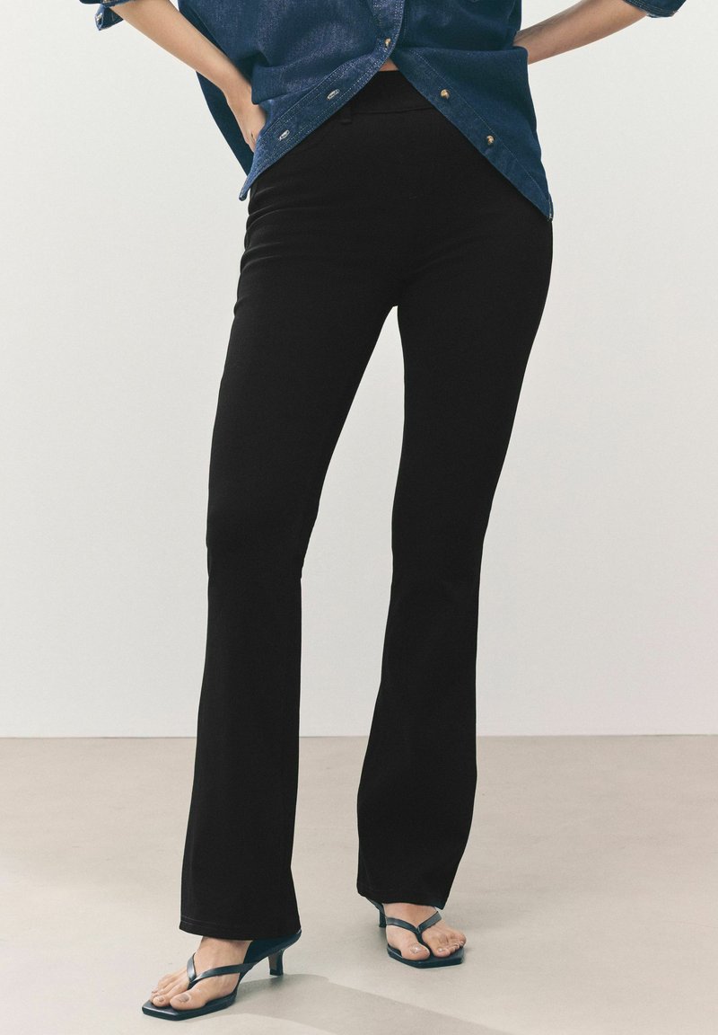 Schwarze, weit geschnittene Hosen mit glatter Textur, hohem Bund und taillierter Passform. Kombiniert mit blauen Flip-Flops und einem Jeanshemd.