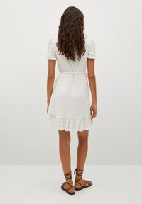 Vestido blanco con mangas cortas abullonadas, cintura ceñida y dobladillo de volantes en capas. Hecho de tela texturada, estilizado con un cinturón atado. Sandalias marrones visibles.