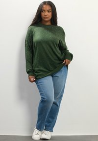 Frau in einem langen grünen Pullover mit kleinen weißen Punkten, blauen Jeans und weißen Sneakern, die vor einer schlichten weißen Wand steht.