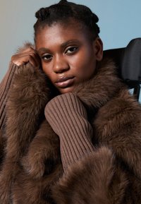 Νεαρή γυναίκα με πλεγμένα μαλλιά, ντυμένη με καφέ γούνινο παλτό και ριγωτά πλεκτά γάντια, στηρίζεται σε μια πλάτη αναπηρικής καρέκλας μπροστά σε ουδέτερο φόντο.