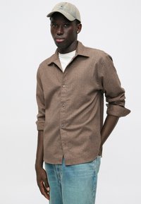 Καφέ μοτίβο μακρυμάνικου πουκάμισου με κουμπί, αναδιπλωμένα μανίκια, συνδυασμένο με ανοιχτό μπλε τζιν και μπεζ καπέλο. Λείος υφασμάτινος ιστός.