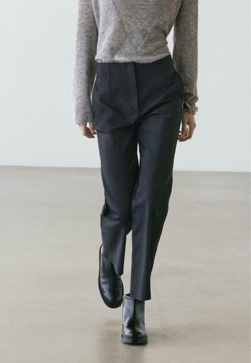 Pantaloni neri su misura con taglio dritto, piega definita e tasche laterali, abbinati a un maglione grigio lavorato a maglia e stivaletti neri.