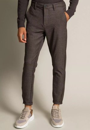 Mężczyzna noszący brązowe, slim-fit w kratę spodnie z mankietami u nogawek oraz beżowe buty sportowe, stojący na jednolitym tle.