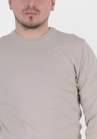 Camicia beige a maniche lunghe in tessuto a coste, con colletto rotondo e un logo a contrasto sul petto. Texture liscia, design aderente.
