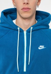 Zilā flīsa kapuce ar pus-izvelkamu dizainu; aprīkota ar piparmētru zaļu rāvējslēdzēju un auklām, kā arī ar baltu Nike logotipu uz krūtīm.