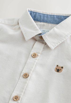 Beiges Baumwollhemd mit einem Chambray-Kragen, Holzknöpfen und einem auf der linken Seite eingestickten Bärengesicht.