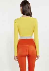 Mouwloze gele crop top met een witte tailleband, gecombineerd met oranje high-waisted leggings met geplooide details aan de achterkant.