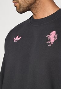 Felpa girocollo in tessuto grigio scuro; presenta il logo Adidas ricamato in rosa e una grafica dettagliata di un leone rosa sul davanti.