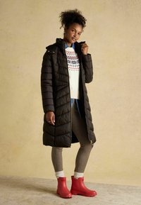 Čierny prešívaný dlhý pufový kabát s vysokým golierom, biely sveter s červeným a modrým vzorom, kockované legíny a červené členkové čižmy.