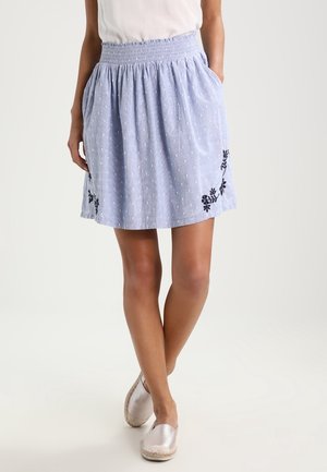 Kobieta nosząca jasnoniebieską spódnicę z marszczonym stanem, subtelną kropkowaną fakturą, czarnym kwiatowym haftem blisko dołu i białymi butami wsuwanymi.