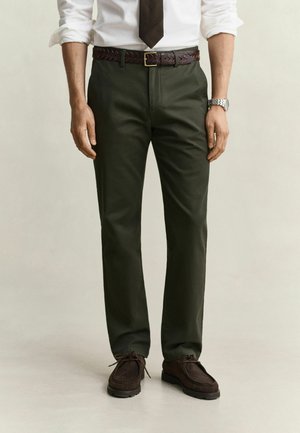 Man draagt donkergroene broek, wit overhemd met opgerolde mouwen, donkere stropdas, gevlochten bruine riem, horloge en donkerbruine veterschoenen.