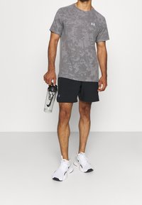 Camisa de manga corta de camuflaje gris, pantalones cortos negros y zapatillas deportivas blancas. Sosteniendo una botella de agua transparente con tapa negra.