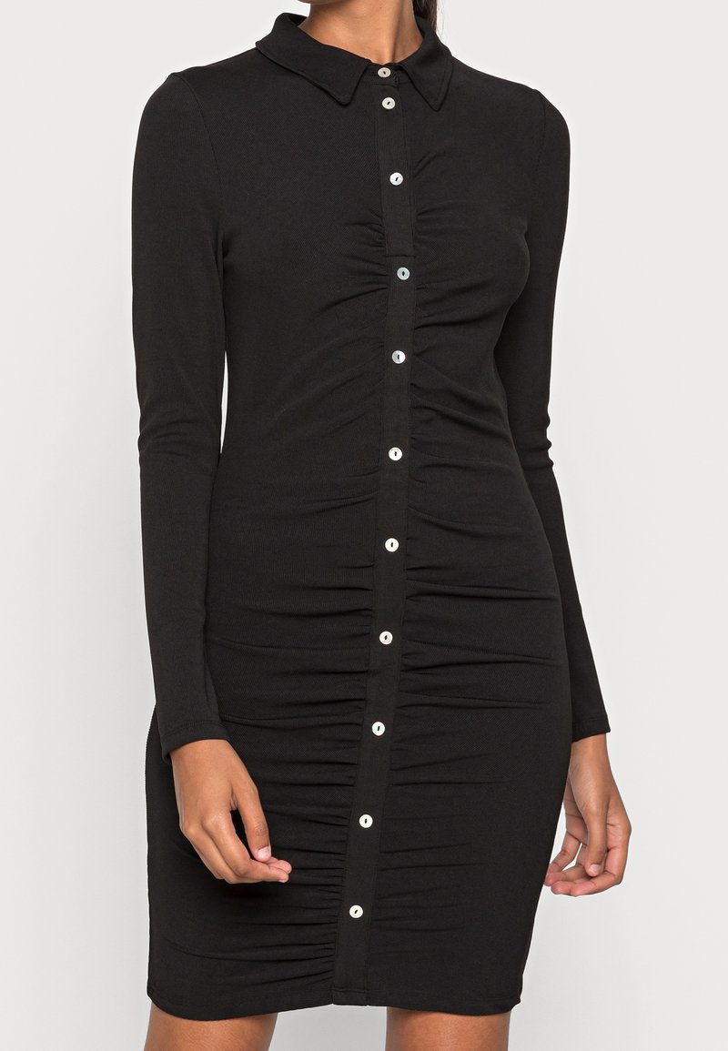 Robe noire à manches longues en tissu côtelé, comportant une ouverture boutonnée à l'avant avec des boutons blancs et une texture plissée le long du corps.