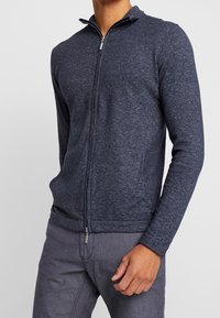 Tmavě modrý zip-up svetr s vysokým límcem, žebrovanými manžetami a bočními kapsami, vyrobený z texturované, lehké látky.