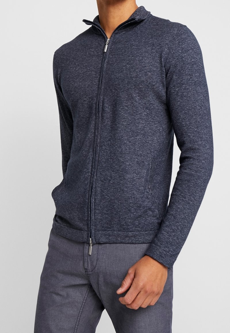 Tmavě modrý zip-up svetr s vysokým límcem, žebrovanými manžetami a bočními kapsami, vyrobený z texturované, lehké látky.