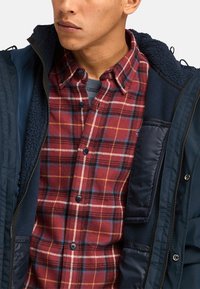 Marineblauwe zip-up jas met een zachte binnenvoering over een rood geruit overhemd met gele en blauwe accenten. Knopsluiting en een zachte textuur.