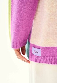 Ruka odpočívajúca pri leme farebne blokovaného svetra v ružovej, krémovej, zelenej a fialovej farbe s štítkom "Naoko Knitwear".