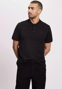 Czarna koszulka polo z bawełny z krótkimi rękawami, zapięcie na trzy guziki oraz minimalnym logo na klatce piersiowej. Noszona z czarnymi spodniami.