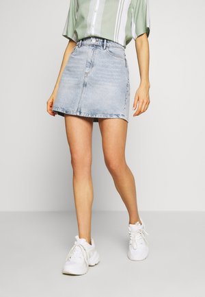 Person trägt ein hellgrün gestreiftes Hemd, hellblauen Denim-Minirock und weiße Sneakers und steht vor einem einfarbigen Hintergrund.