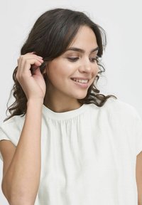 Ljusfärgad, kortärmad topp med en samlad halsringning. Tillverkad av ett mjukt, strukturerat tyg. Modellen har lockigt hår och handen i håret.