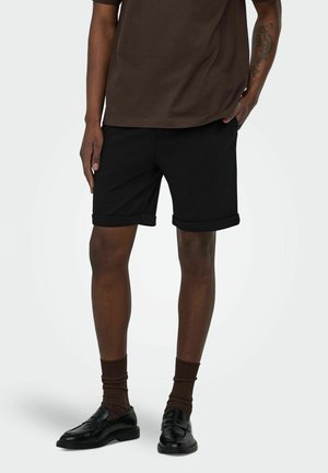 Persona con pantalones cortos negros remangados, calcetines marrones, mocasines negros y una camisa marrón de manga corta con una mano en el bolsillo.
