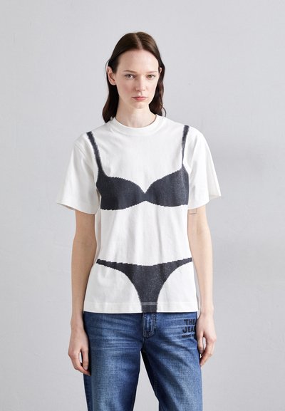 Λευκό βαμβακερό T-shirt με μαύρη γραφική εκτύπωση ενός στηθόδεσμου και μίας εσώρουχο. Χαλαρή εφαρμογή με κοντά μανίκια και στρογγυλή λαιμόκοψη. Φορεμένες μπλε τζιν παντελόνες.