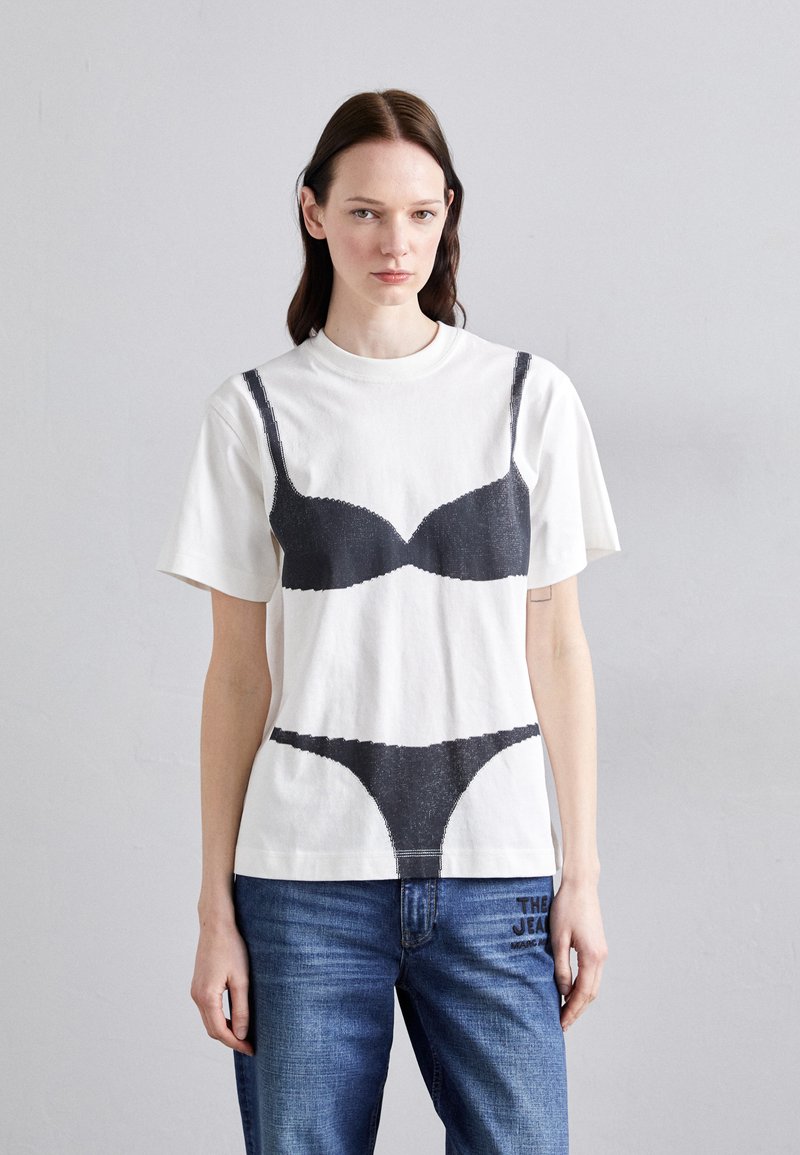 White cotton T-shirt featuring a black graphic print of a bra and thong. Relaxed fit with short sleeves and a round neckline. Blue denim jeans worn.