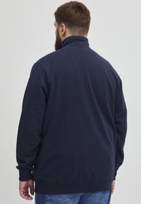 Veste zippée bleu marine foncé en tissu doux. Elle présente un col montant et des poignets côtelés, avec une finition lisse et texturée.