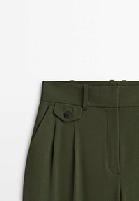 Pantalon taille haute vert olive avec passants de ceinture, rabat de poche côté gauche fixé par un bouton noir, et plis à l'avant.
