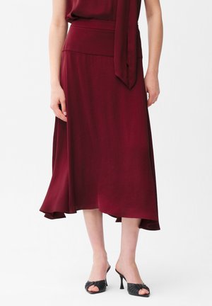 Frau trägt einen burgunderroten Midirock mit asymmetrischem Saum und einem passenden Oberteil, kombiniert mit schwarzen, offenen Sandalen mit Absatz.