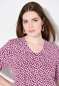 Blusa morada con un patrón floral, que presenta pequeñas margaritas blancas. Tiene mangas cortas, un escote redondeado y detalles de pliegues.
