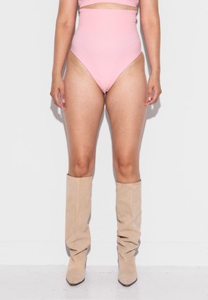 Kobieta w różowych majtkach od bikini z wysokim stanem i beżowych zamszowych kozakach sięgających kolan, stojąca przy białej ścianie.