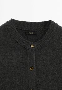 Cardigan tricoté gris foncé avec un col rond et une fermeture à bouton en forme de nœud doré. Texture douce et design épuré sans motifs visibles.