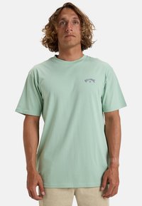 Licht mintgroene katoenen T-shirt met korte mouwen, ronde hals en een klein paars grafisch logo op de borst. Zachte textuur, ontspannen pasvorm.