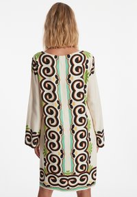 Langermige jurk in crème met zwarte, groene en oranje abstracte patronen. Beschikt over een ronde halslijn en een middel lange zoom. Glad textuur van de stof.