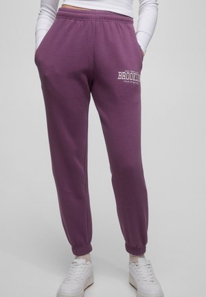 Personne portant un pantalon de jogging violet avec le texte "Brooklyn" sur la cuisse, associé à des baskets blanches et un haut à manches longues blanc.