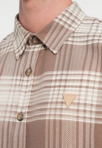 Brun og krem rutete skjorte med knapp-ned krage, med tre-lignende knapper og et trekantet lærmerke på venstre bryst.