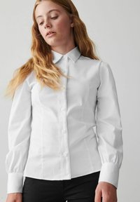 Camicia bianca a maniche lunghe con colletto button-down, patta frontale e maniche a sbuffo. Realizzata in un tessuto liscio con una vestibilità strutturata.