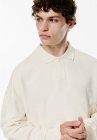 Polo de culoare crem, cu o textură moale, având un guler cu două butoane și mâneci relaxate. Design simplu, fără modele sau accesorii.