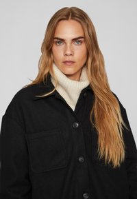 Črna majica-jakna iz volnene mešanice z gumbnim zapiranjem, velikim žepom in bež pletenim rolkom spodaj, z gladko teksturo in prilegajočim krojem.