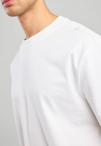 Valkoinen pyöreäkauluksinen collegepaita, joka on valmistettu pehmeästä puuvillasta. Paidassa on rento leikkaus, hienovaraiset ommellut yksityiskohdat ja sileä pinta.